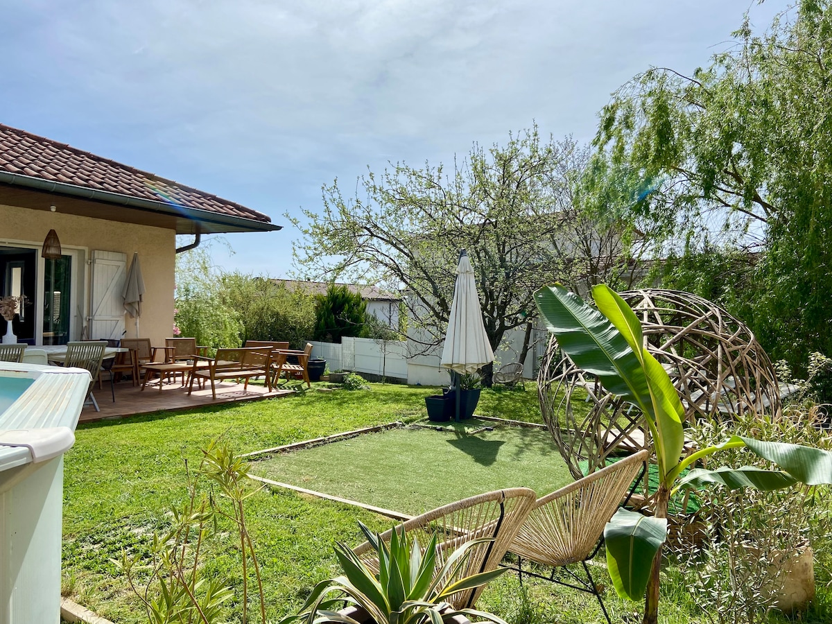 An outdoor area features a well-maintained garden with green grass and various plants. A patio with garden furniture and a large umbrella provides shaded seating. A wooden structure in the background complements the serene setting.