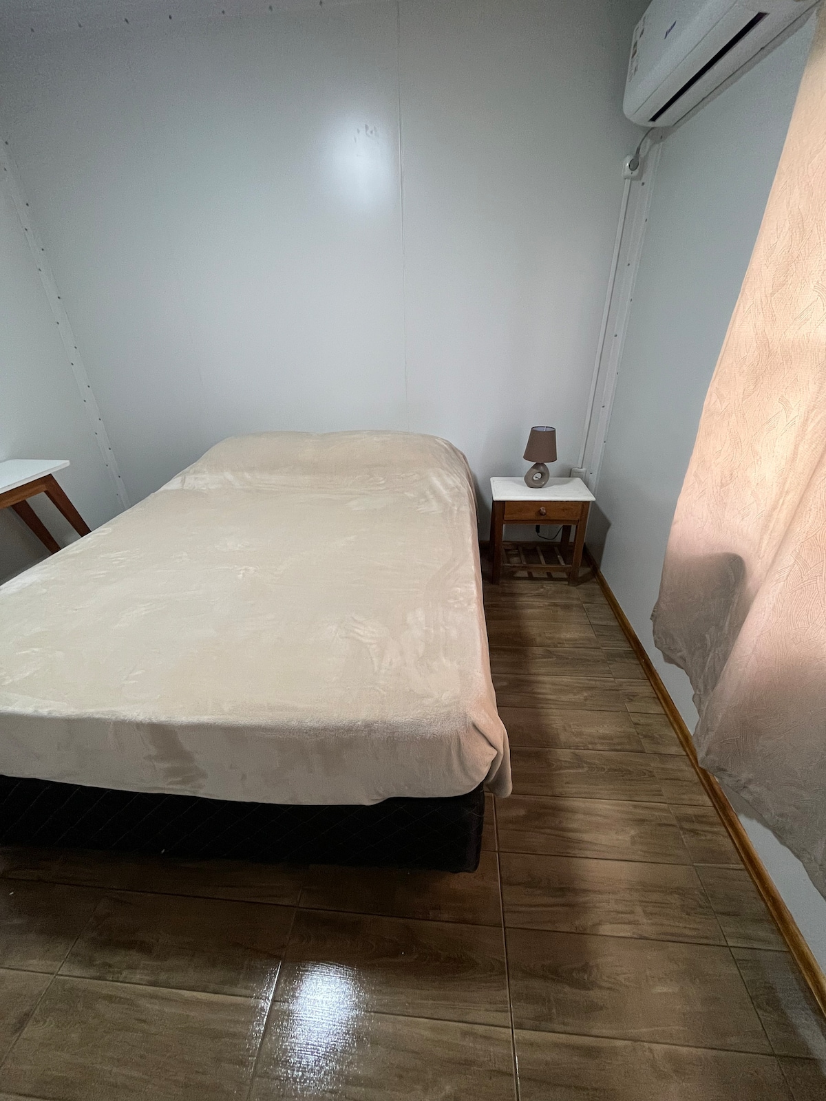 A simple bedroom features a spacious bed covered with a neutral blanket. A nightstand with a lamp is positioned beside the bed, and a window partially draped with sheer curtains allows natural light to enter. The floor is tiled, enhancing the room's clean aesthetic.