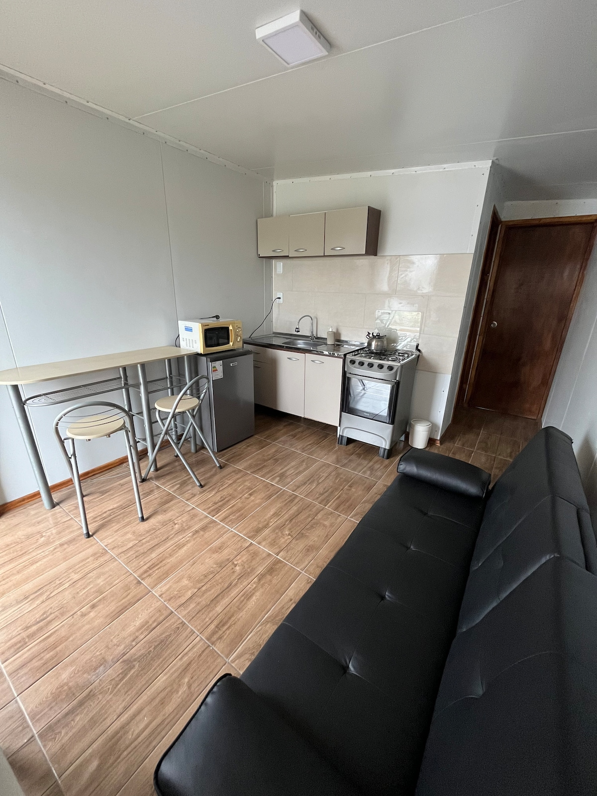A compact kitchen area features modern cabinetry and sleek countertops. A microwave sits over a small refrigerator, while a stovetop oven is nearby. Two stools are positioned at a narrow table, and a black sofa is visible in the seating area, completing the contemporary layout.