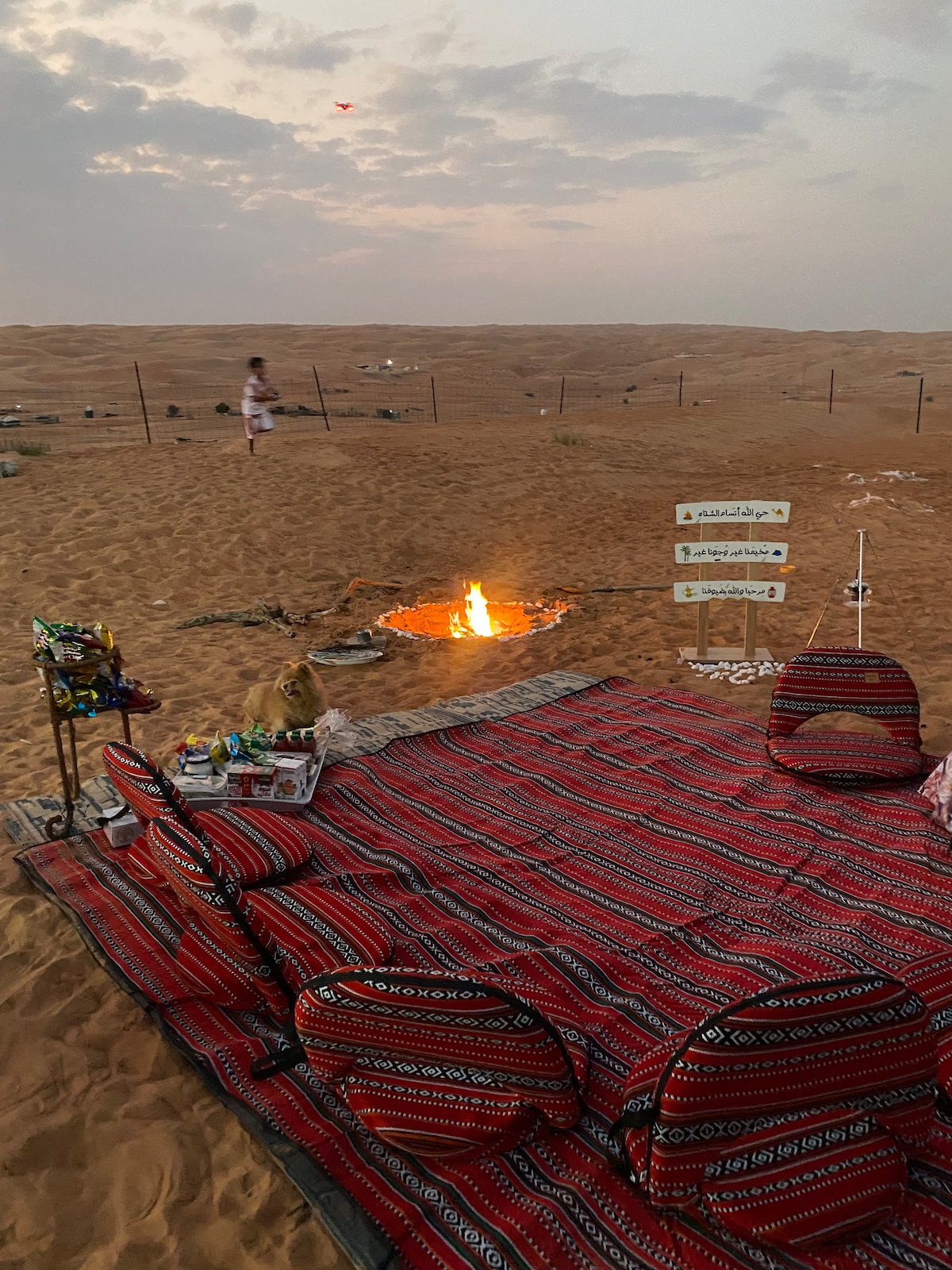A vibrant red and black patterned carpet is laid on the sand, surrounded by low seating cushions. A flickering fire is positioned nearby, creating a warm glow against the soft desert background. Distant sand dunes are visible under a partly cloudy sky.