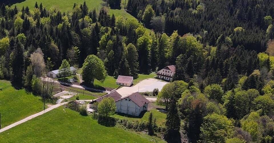 Aerial view of a serene natural landscape featuring lush green fields and dense forests. Three distinct buildings are visible, each with a varying roof style, surrounded by trees. A winding path and well-maintained outdoor spaces connect the structures peacefully.