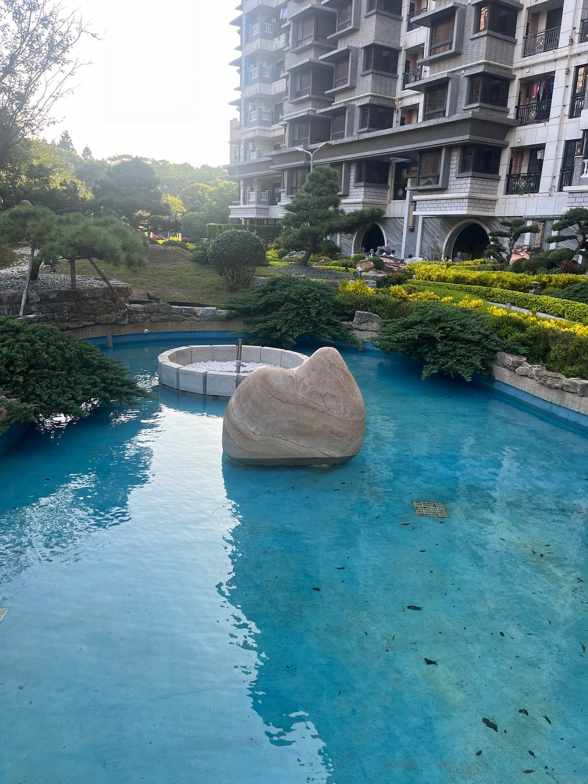 A serene outdoor area features a clear blue pond, with a large smooth stone placed centrally. Lush greenery and flower beds surround the water, complemented by a multi-story building in the background. A subtle fountain can be seen in the pond, offering a calming presence.