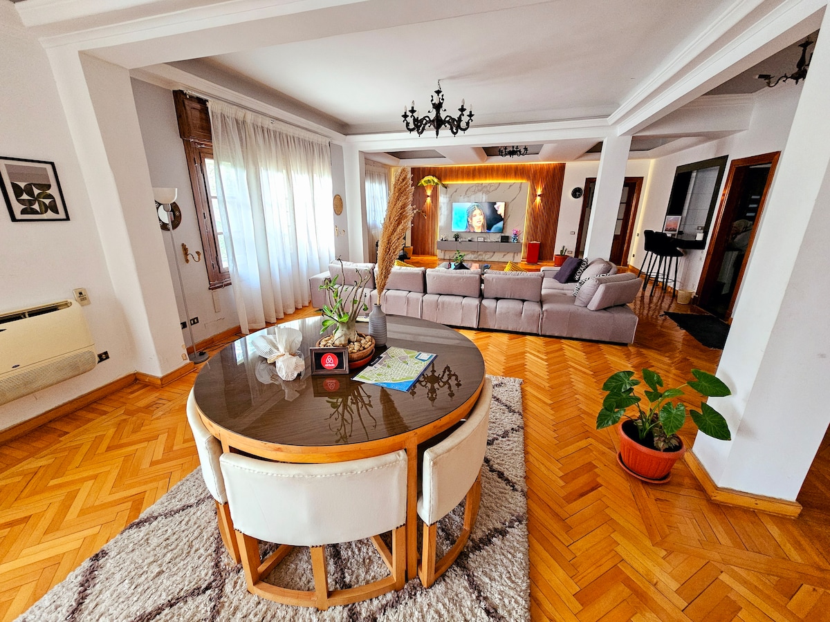 The spacious living area features a circular dining table surrounded by four light-toned chairs. A large, comfortable sectional sofa is arranged nearby, accented by soft cushions. Natural light filters through sheer drapes, illuminating the warm wooden flooring and modern decor, creating a welcoming ambiance.