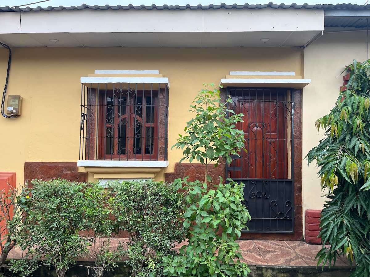 The exterior features a charming façade with a blend of warm colors. Two windows are adorned with decorative metal grills, and a wooden door is framed by a wrought-iron gate. Lush greenery surrounds the entrance, creating a welcoming environment.