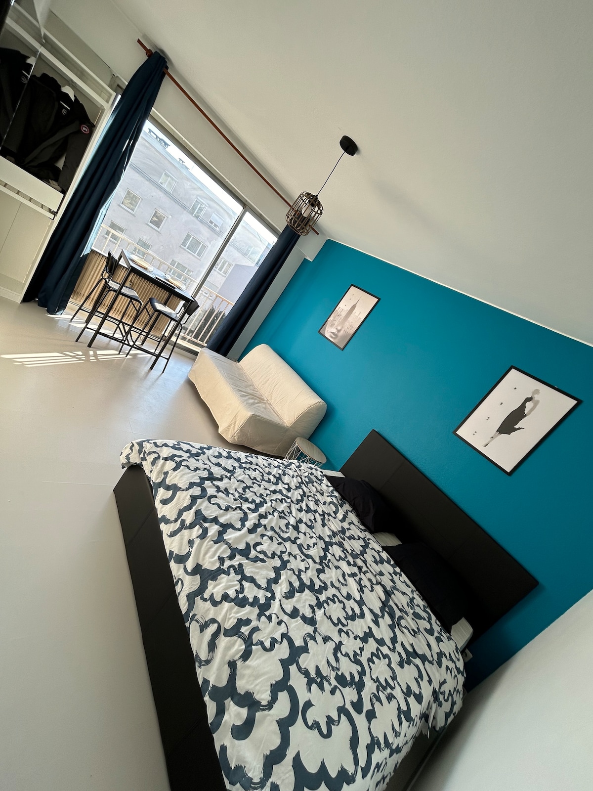 A spacious studio features a large bed adorned with a patterned duvet, complemented by a cozy sofa bed. A bold blue accent wall creates a contemporary look, while natural light illuminates the space through a large window with sheer curtains. A small dining table is visible near the balcony.