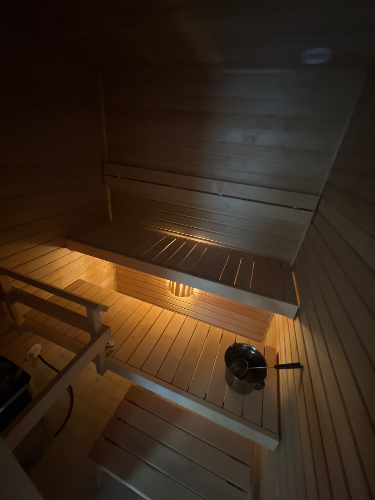 A cozy wooden sauna interior is illuminated by soft lighting, highlighting the slatted benches that offer ample seating. A steaming kettle sits on a lower shelf, adding to the tranquil ambiance of the space designed for relaxation.