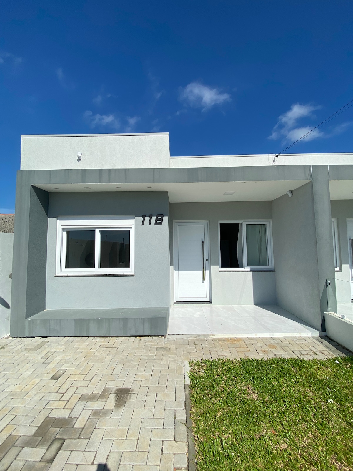 The exterior of the house is highlighted by a modern design with light grey walls and large windows. A neatly paved path leads to the front door, which is framed by a small lawn area. Clear blue skies add to the inviting atmosphere.