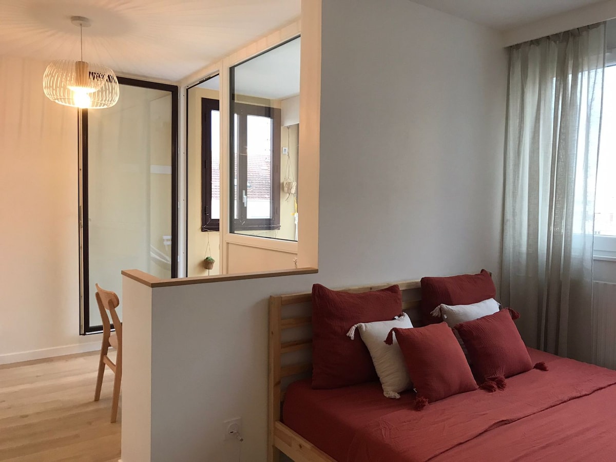 A spacious sleeping area features a wooden bed adorned with red decorative pillows. Natural light enters through a large window, and a modern light fixture hangs from the ceiling. Glass partitions create separation between the bed and a small workspace in the background.