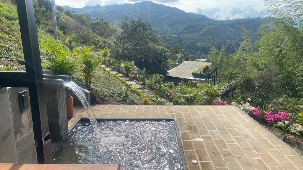 A private outdoor area features a small hot tub with a gentle water flow, surrounded by a stone patio. Lush greenery lines the space, with vibrant flowers adding color. The view includes rolling hills and distant mountains under a sunny sky.
