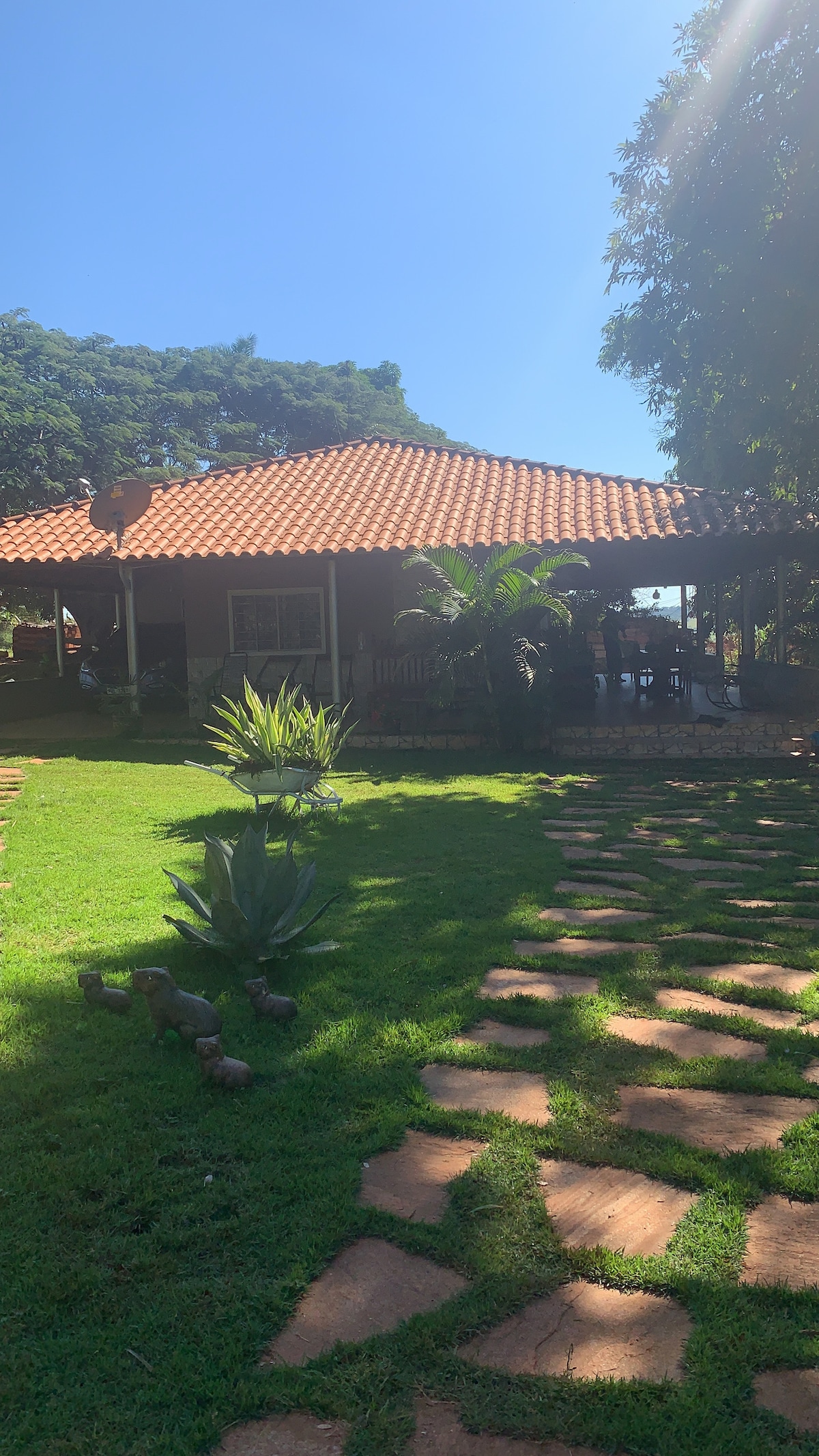A charming house with a terracotta-tiled roof is set against a clear blue sky. Lush greenery surrounds the building, with a stone pathway leading to the entrance. The outdoor area features well-maintained grass, providing a serene ambiance.