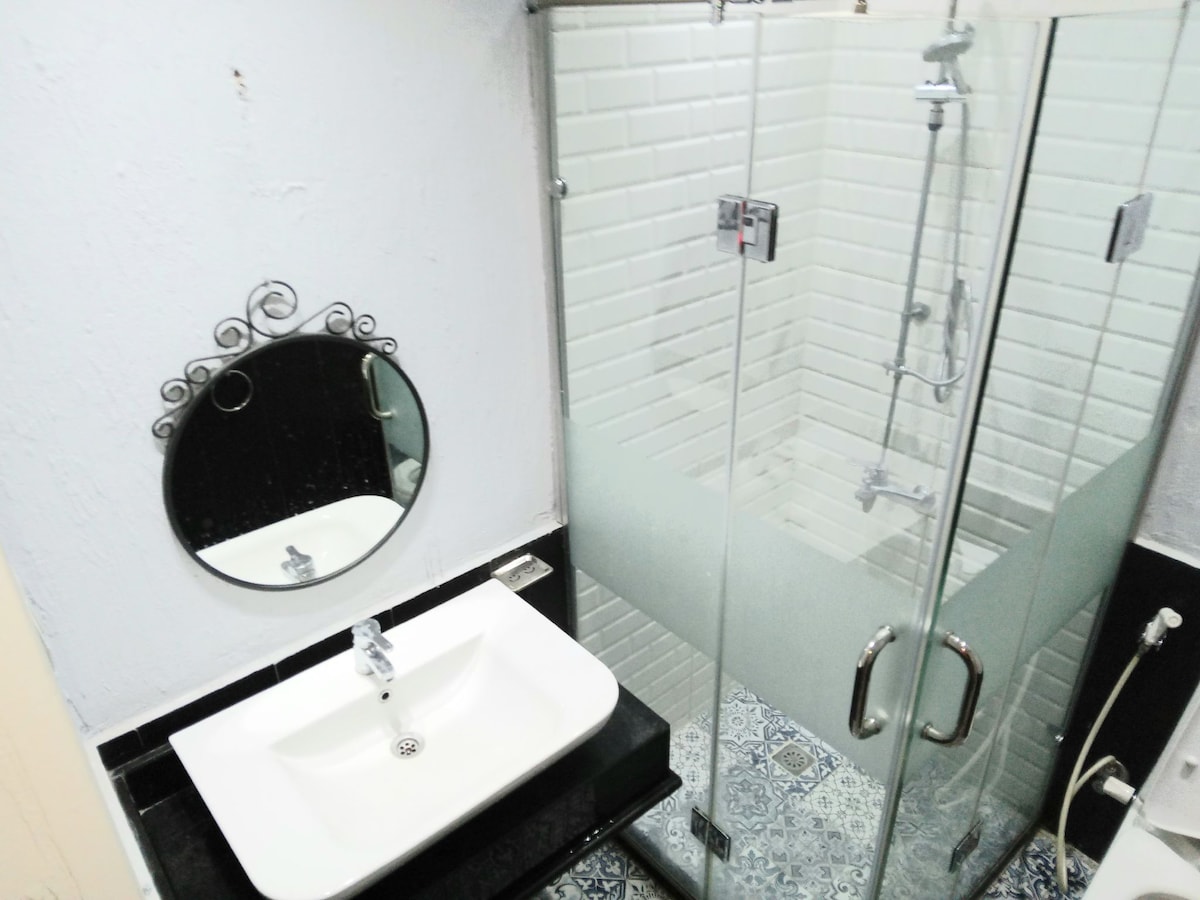 The bathroom features a modern glass shower enclosed by frosted panels, complemented by stylish tiled walls. A sleek, white sink with a chrome faucet is positioned on a dark countertop, while a round mirror hangs above, enhancing the space's contemporary design.