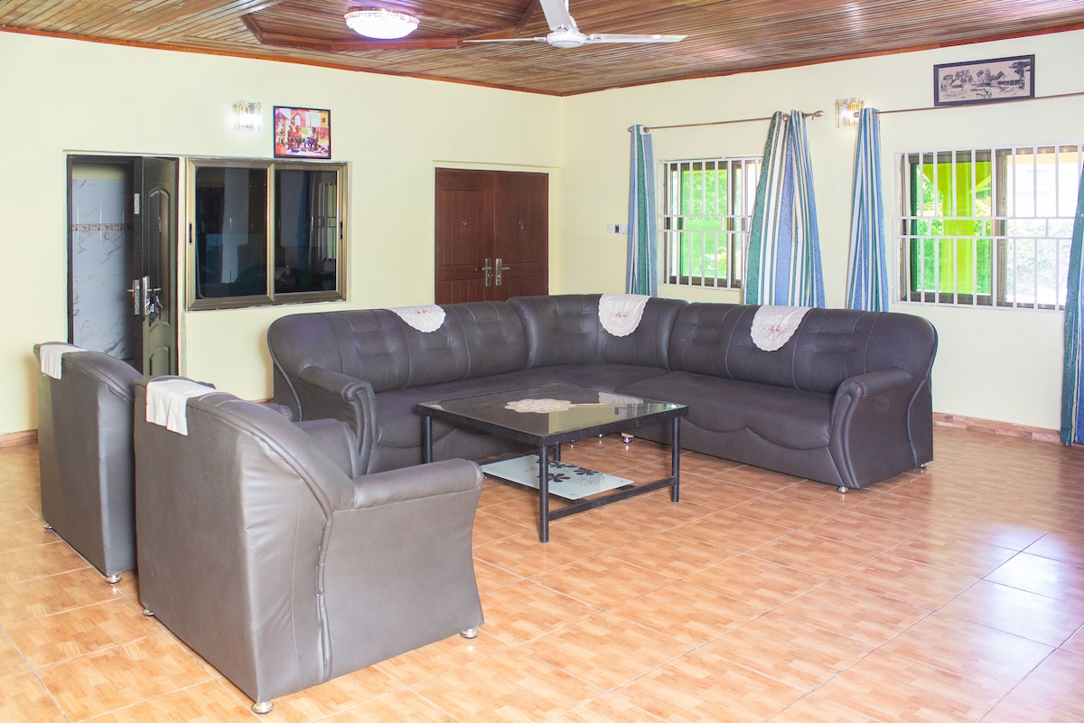 A spacious living area is furnished with a large sectional sofa and two armchairs, arranged around a central coffee table. Large windows with striped curtains allow natural light to filter in, while a ceiling fan provides ventilation. Double doors lead to another room.