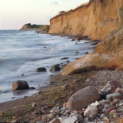 A rugged coastline is depicted, featuring a sandy beach with scattered smooth stones. The cliffs rise in the background, showcasing layers of earth. Gentle waves lap at the shore, while the sky transitions to soft hues, creating a serene natural setting.