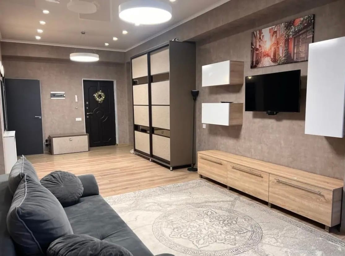 A spacious living area features a comfortable sofa in front of a modern entertainment unit. Light wood furniture complements the neutral-toned walls, while decorative elements like wall art enhance the welcoming atmosphere. Large ceiling lights illuminate the open space, creating a warm environment.