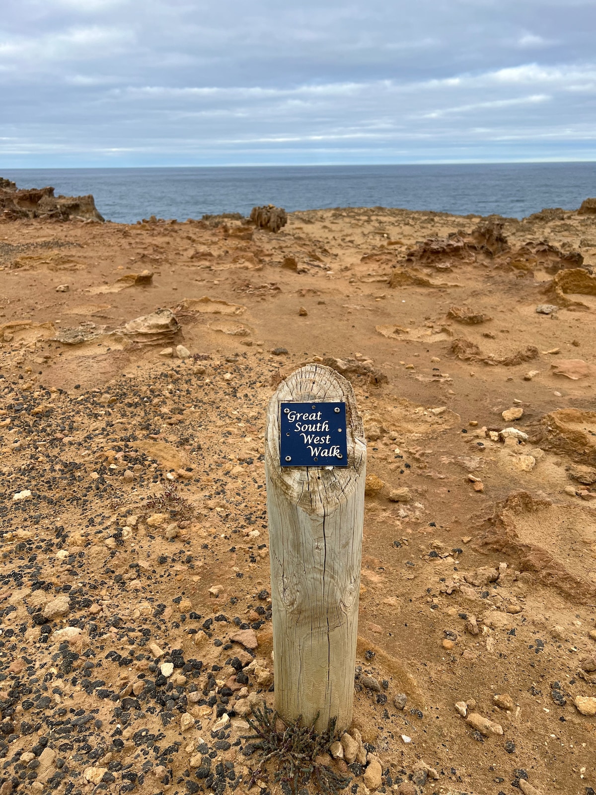 A weathered wooden signpost marks the starting point of the Great South West Walk. The surrounding landscape features rocky terrain and sandy ground leading towards the expansive ocean, under a cloudy sky. The horizon is visually split between the land and water.