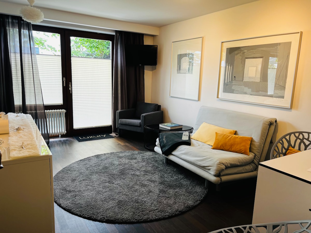 A cozy living area features a spacious gray rug centered under an adjustable couch, complemented by a yellow accent pillow. Natural light filters through sliding glass doors, framed by dark curtains, while a comfortable chair and a wall-mounted television complete the inviting space.