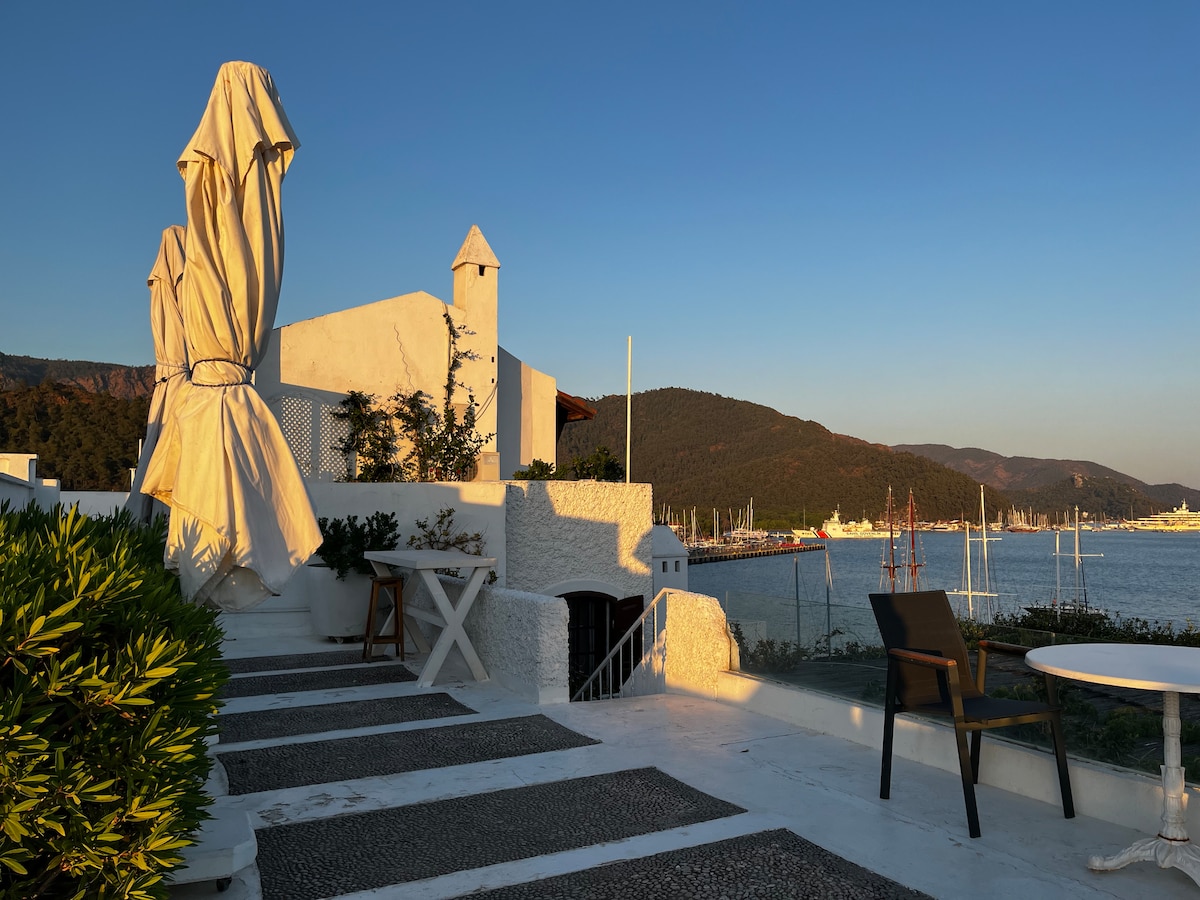 A serene outdoor area features a white building overlooking the water, surrounded by lush greenery. A large, unfurled umbrella stands beside a dining table and chairs, while steps lead down to the shoreline. Mountains are visible in the background under a clear sky.