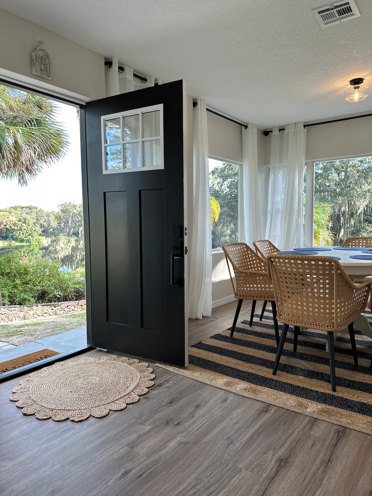 An open door reveals a well-lit dining area featuring a table surrounded by four woven chairs. Large windows provide views of the lush greenery outside, while sheer curtains allow natural light to filter in. A round natural fiber rug lies on the hardwood floor near the entrance.