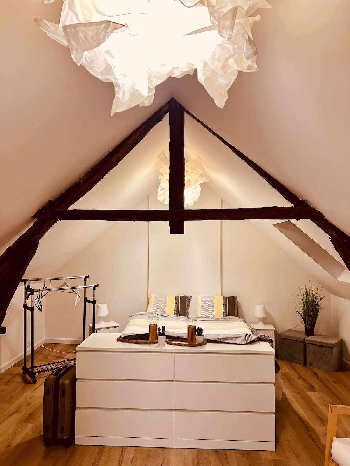 The bedroom features a spacious bed with a light-colored duvet and decorative pillows. A dresser is positioned at the foot of the bed, accompanied by a clothing rack and two suitcases nearby. A large window allows natural light to fill the room, highlighting the wood-beamed ceiling.