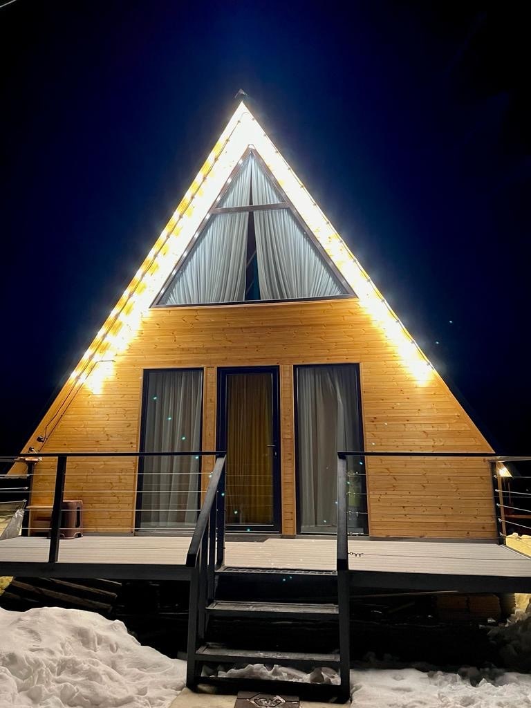 An A-frame cabin is illuminated by string lights, highlighting its wooden exterior against the night sky. Large glass doors are framed by draped curtains, providing a glimpse into the interior. A front deck is visible, surrounded by a snowy landscape.