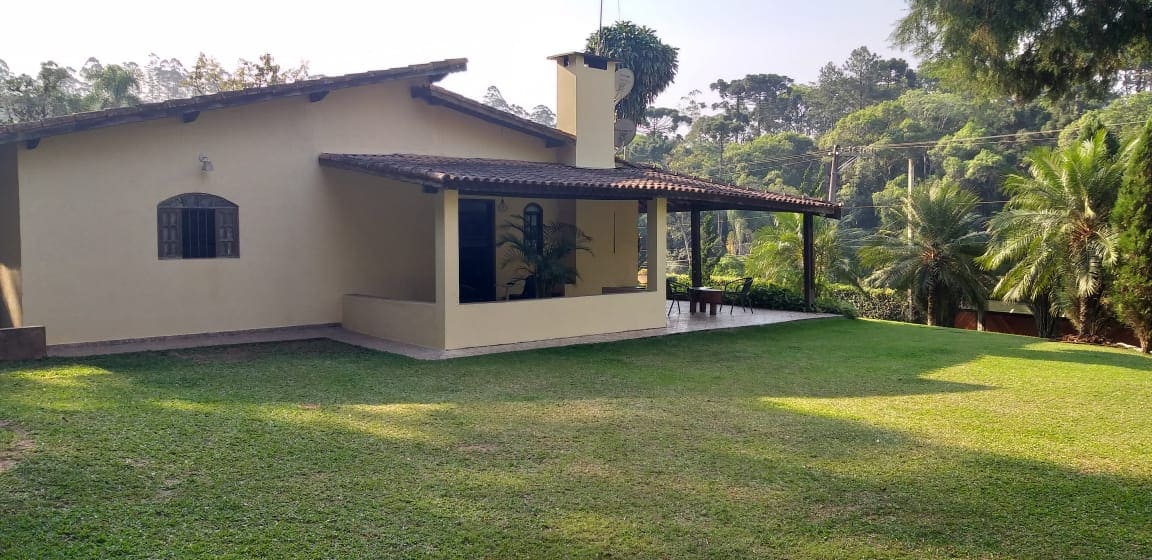 A spacious house with a sloped roof is set against a lush green landscape. A covered porch extends from the front, supported by sturdy columns. The surrounding lawn is well-maintained, bordered by a variety of trees and plants, creating a serene outdoor environment.