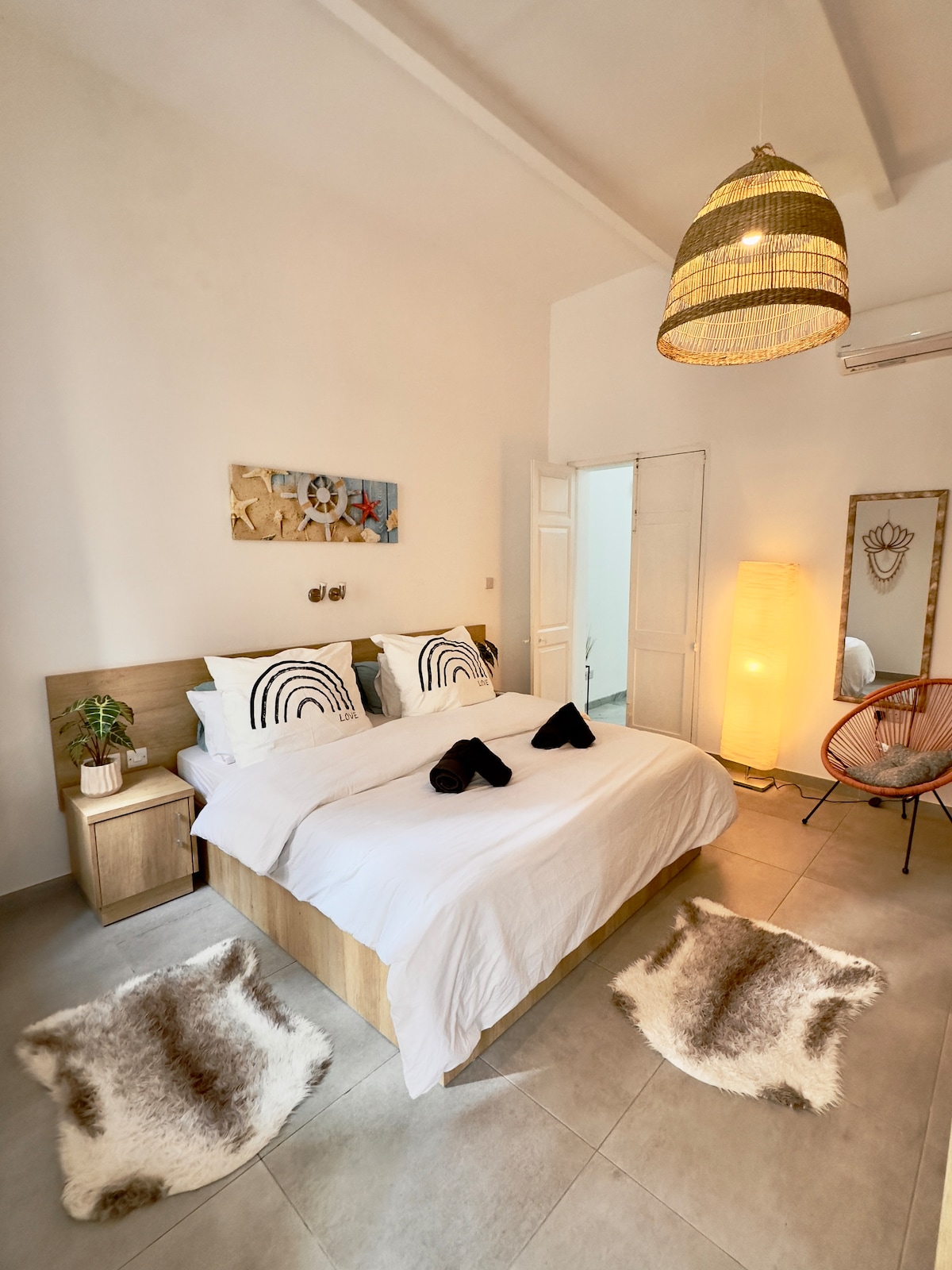 A well-appointed bedroom features a spacious bed with crisp white linens. Decorative cushions add a touch of style, while soft rugs lie on the floor. A woven pendant light casts a warm glow, and a minimalist lamp stands beside the bed, enhancing the serene ambiance.