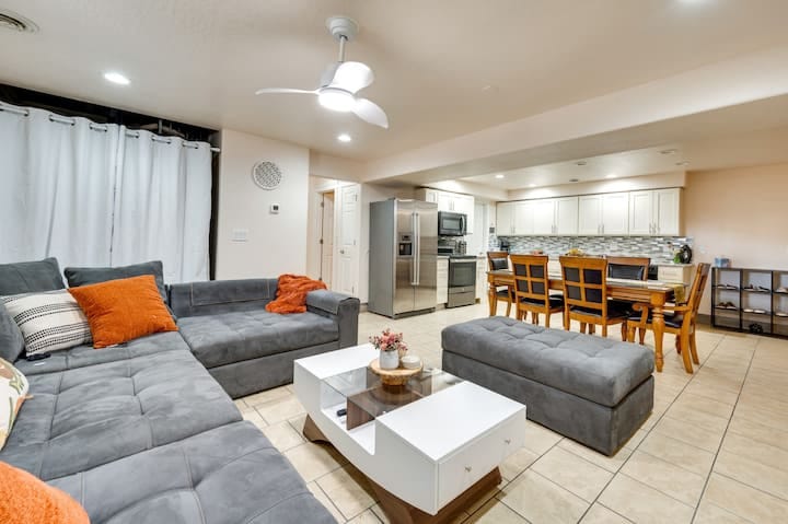 A spacious living area features a large sectional sofa in grey with orange and patterned throw pillows. A central coffee table sits on tiled flooring, accompanied by a dining area with wooden chairs. A light ceiling fan contributes to the airy atmosphere, along with bright ambient lighting.