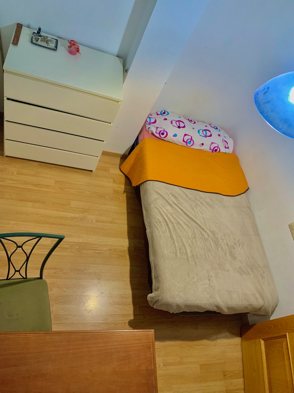 A single bed is positioned against the wall, topped with a colorful blanket and decorative pillows. A small desk and chair are present, alongside a light wood dresser with four drawers. A round ceiling light provides illumination, and warm wood flooring is visible throughout.