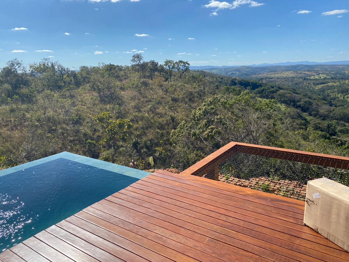 An outdoor deck with wooden flooring overlooks a serene natural landscape. A serene infinity pool seamlessly blends into the horizon, surrounded by greenery. The sky is clear, showcasing vibrant blue with scattered clouds.