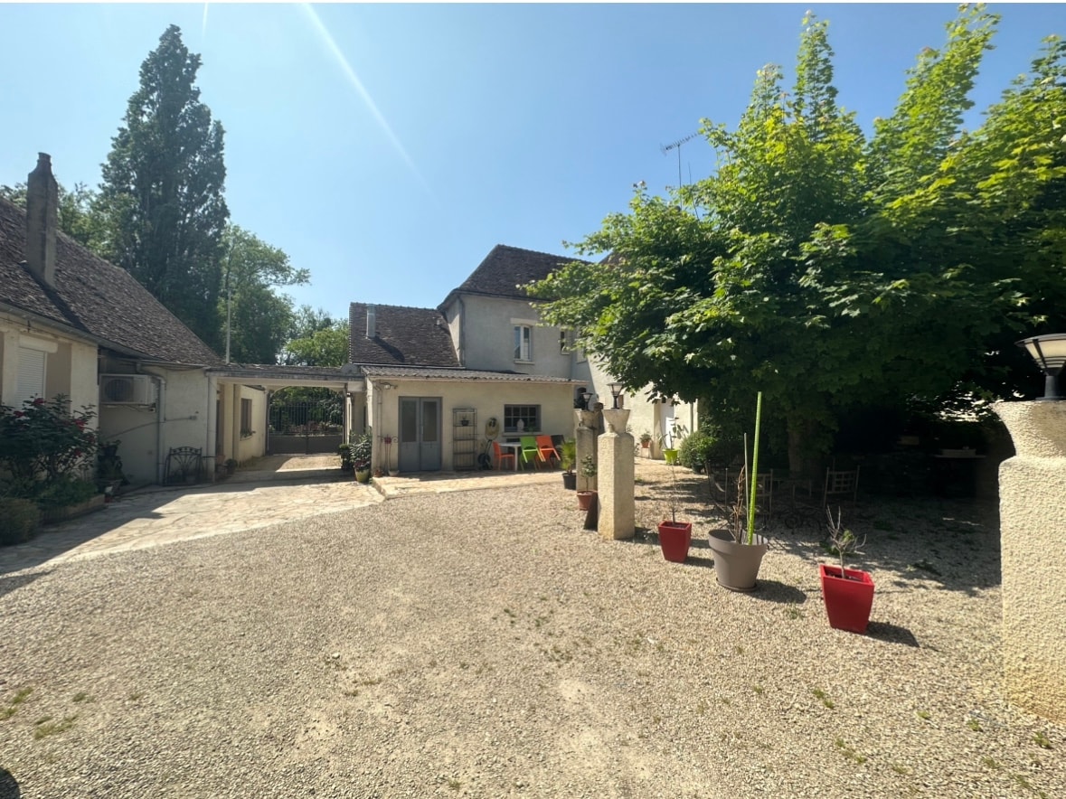 The exterior of the property is shown, featuring a gravel driveway and a landscaped area with various plants and trees. A spacious structure is visible, with a mix of light-colored walls and a traditional roof. Colorful chairs are placed nearby, inviting relaxation.