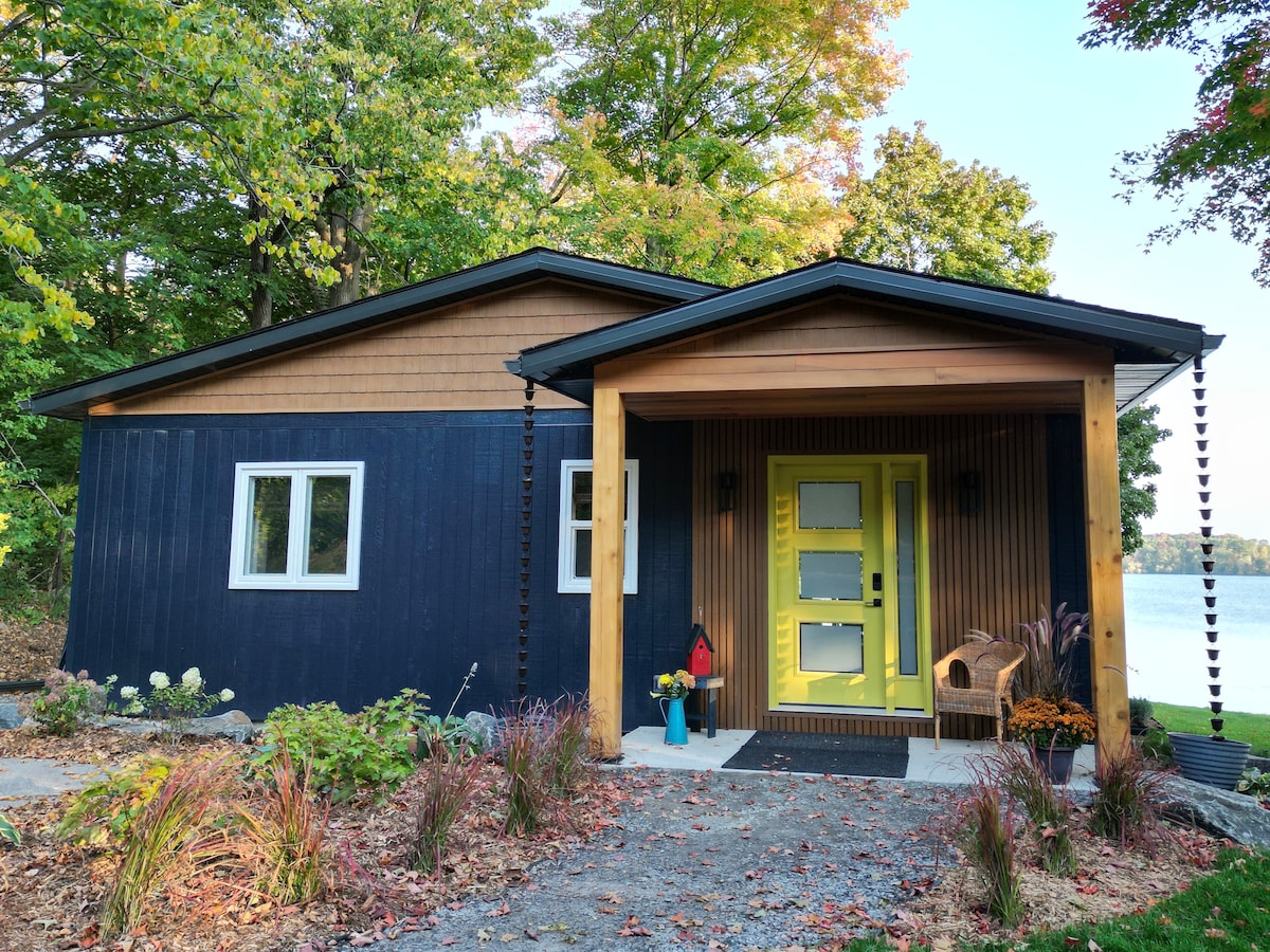 The exterior showcases a contemporary cottage design with dark blue and wooden paneling. A vibrant yellow front door stands prominently, complemented by two side windows. The landscape features various plants and a gravel path leading to the entrance, with the tranquil lake visible in the background.