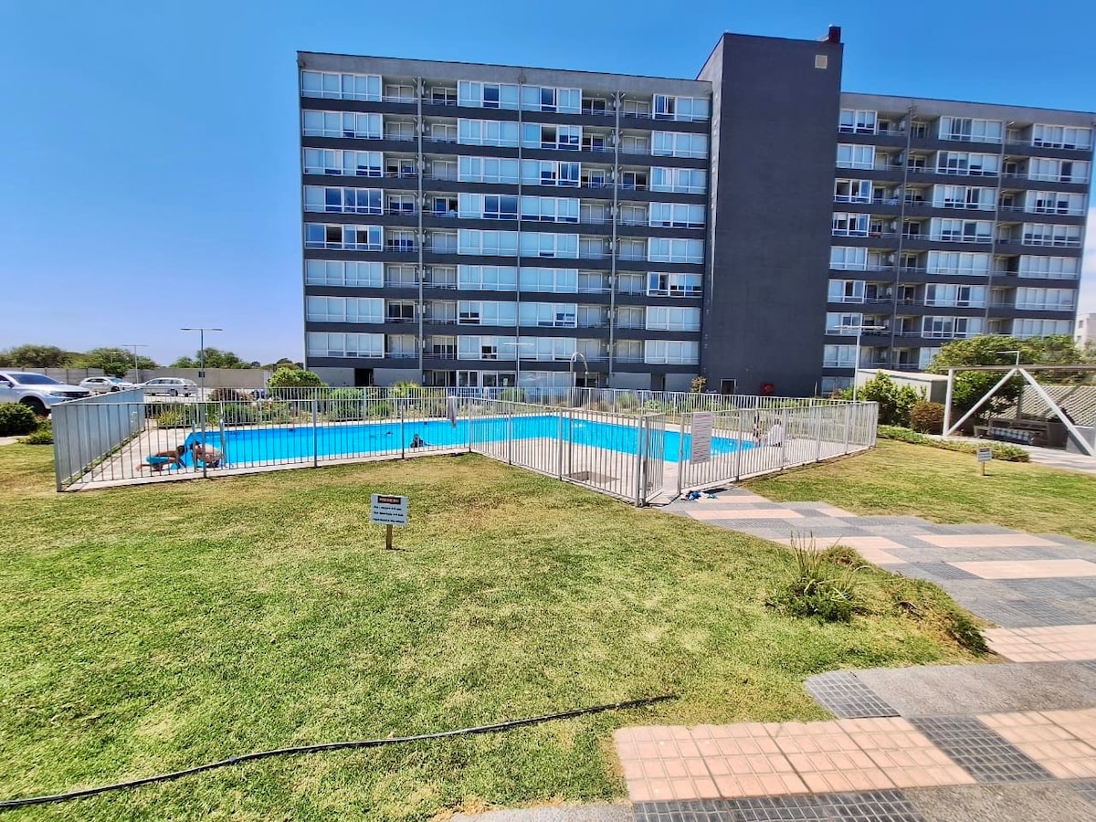 A swimming pool is surrounded by a secure fence, set within a grassy area. The multi-story building features numerous large windows, allowing natural light to brighten the balconies above. Sun loungers are visible near the pool, offering a space for relaxation.