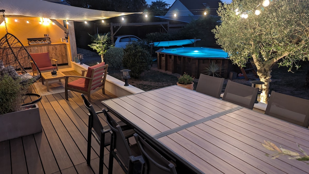 An outdoor terrace is illuminated by string lights, creating a relaxed ambiance. A large dining table is surrounded by several chairs. Nearby, a lounge area features a hanging chair and a wooden deck. A pool is visible in the background, framed by greenery.