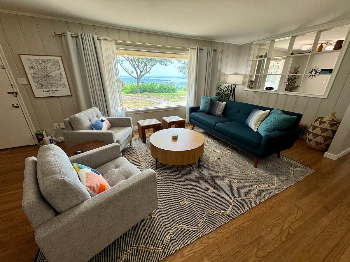 The living area features a modern design with a mint green sofa and two light grey armchairs set around a round wooden coffee table. Natural light floods the space through large windows, offering views of the yard outside. A patterned area rug adds warmth to the wooden floor.