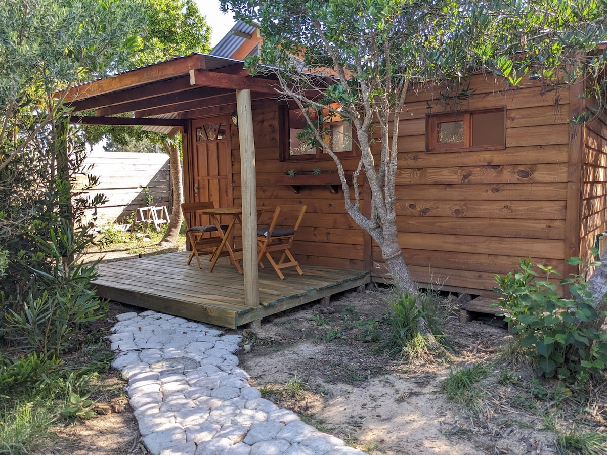 A wooden cabin is showcased with a small deck area featuring two folding chairs and a table. A pathway composed of stones leads up to the entrance, surrounded by lush greenery and a tree, enhancing the natural setting.