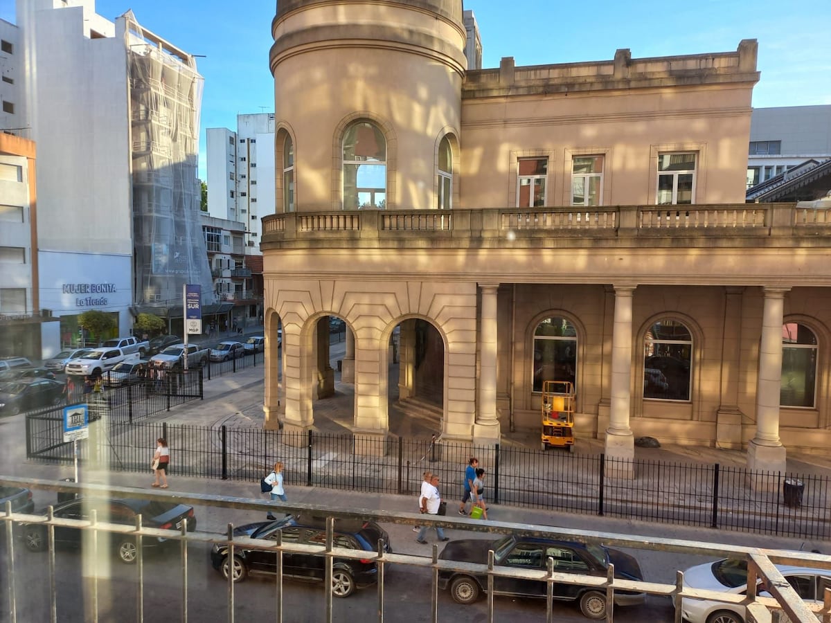 A view of a historic building features arched windows and a rounded tower, framed by a busy street. Pedestrians can be seen walking along the sidewalk, while parked cars line the street. Daylight brightens the scene, highlighting the architectural details.