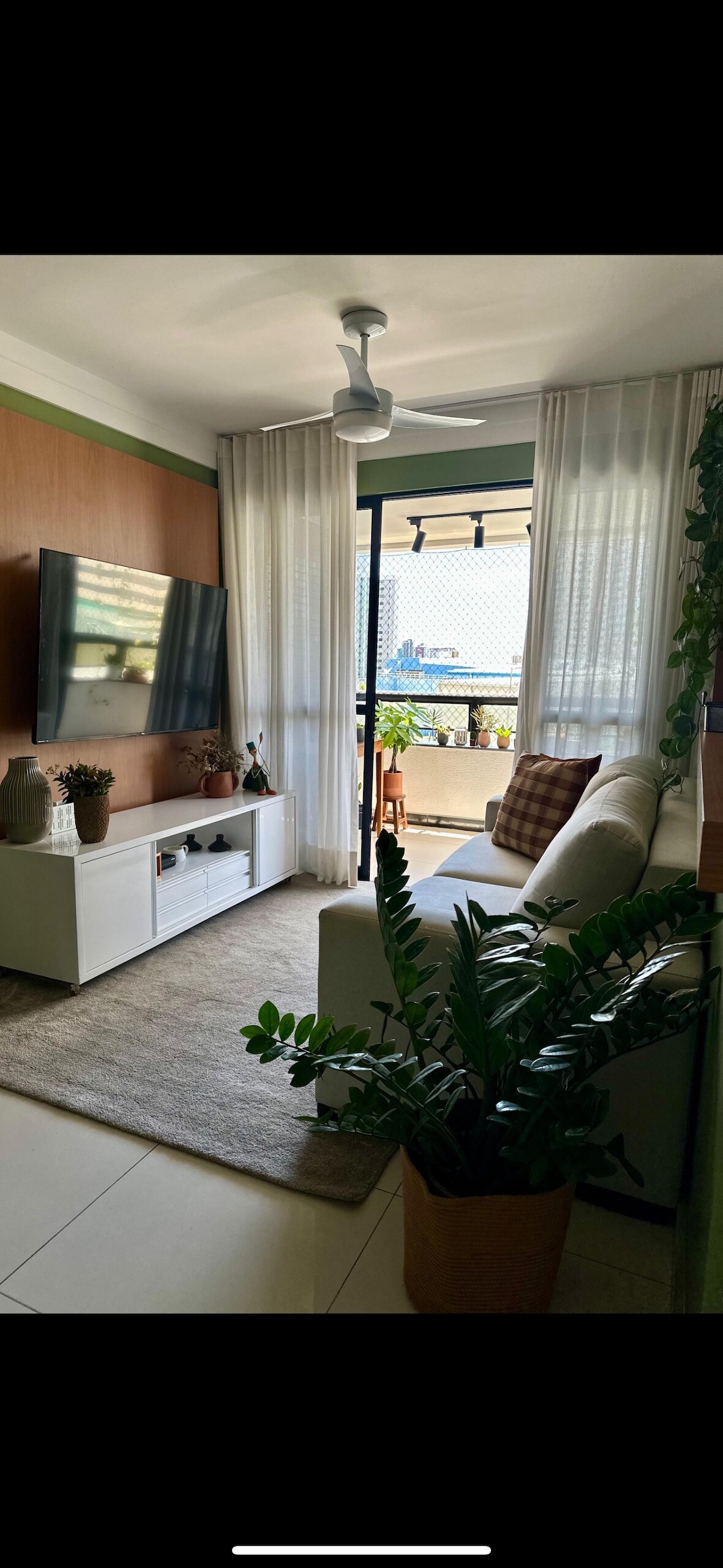 A well-lit living area features a comfortable sofa adorned with a plaid cushion. A television is mounted on a wooden panel, complementing a white media unit. Large glass doors lead to a balcony, with flowing sheer curtains adding a touch of softness to the space.