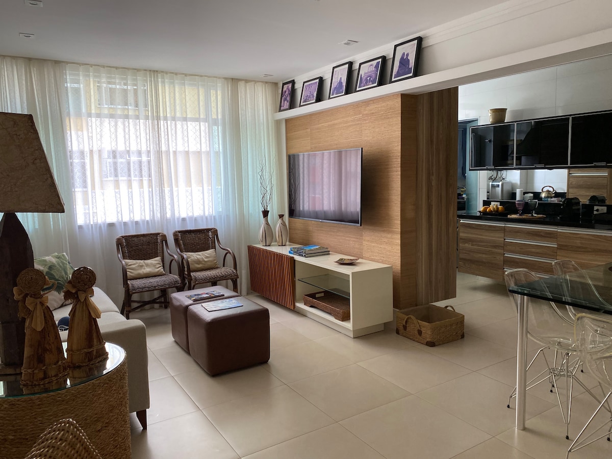 The living area is highlighted by large windows with sheer curtains, allowing natural light to enhance the space. A television is mounted on a wooden wall, and a mix of seating, including two wicker chairs and a beige sofa, provides comfort. Decorative items and a stylish coffee table contribute to the ambiance.