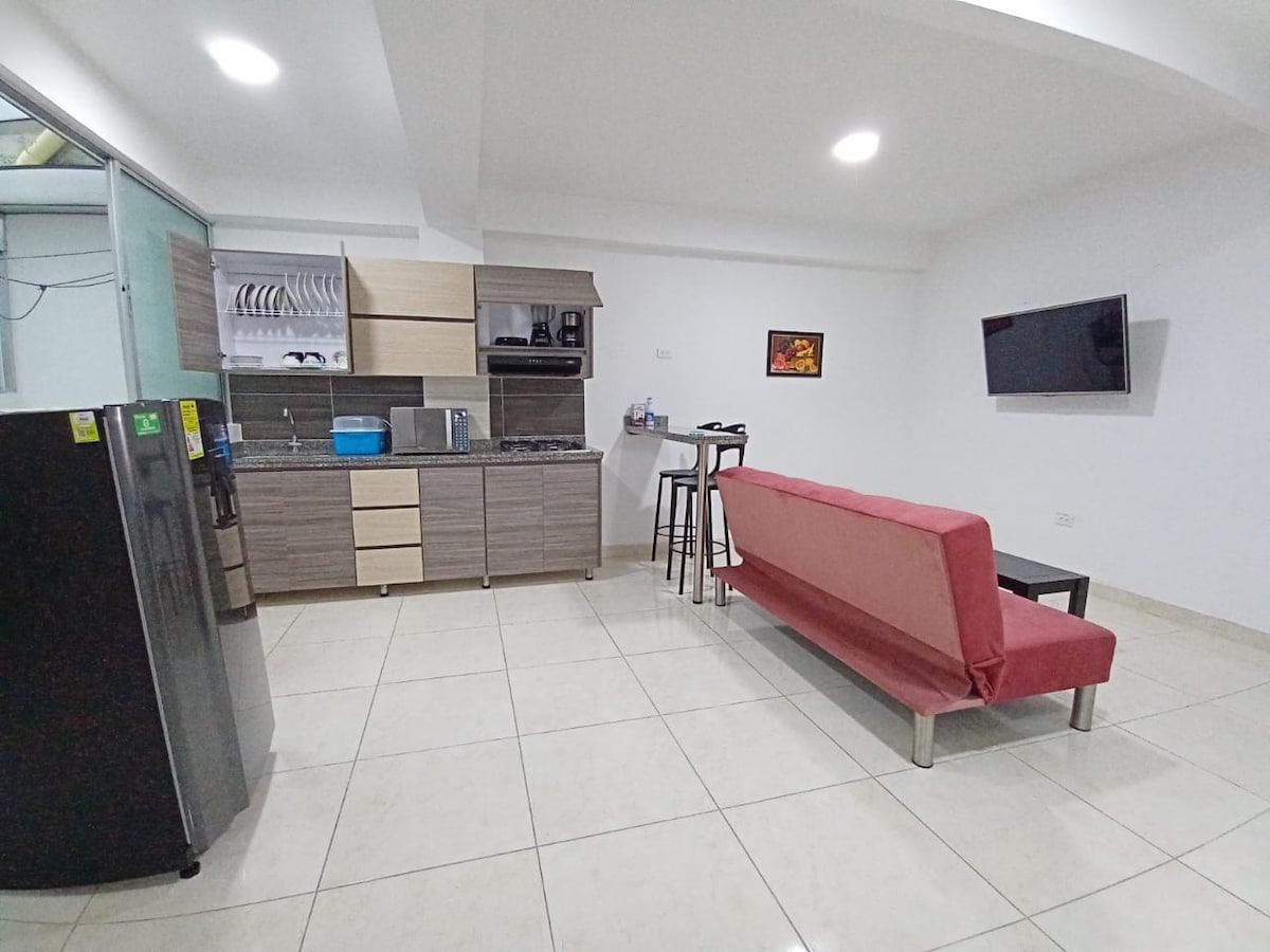 A modern kitchen area is equipped with a refrigerator, stove, and sink, complemented by wooden cabinetry. A stylish dining space features high stools beside a countertop. A sofa in a warm hue sits across from a mounted television, with natural light illuminating the tiled floor.