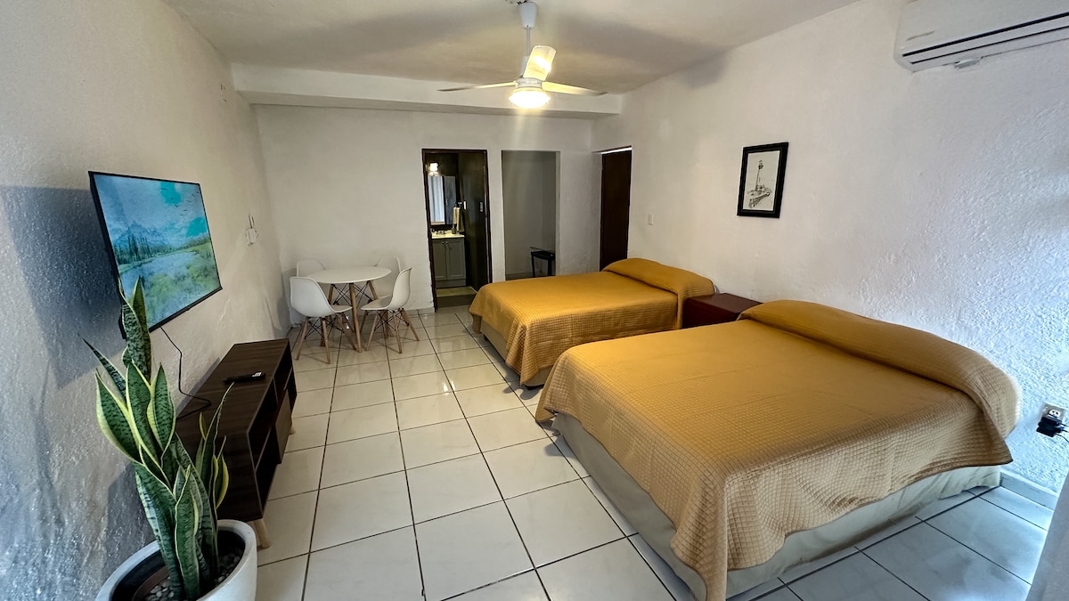 A spacious room is featured with two oversized beds covered in golden-hued linens. A flat-screen television is mounted on the wall opposite. A small dining table with white chairs is visible to one side, while a large plant adds a natural touch to the decor.