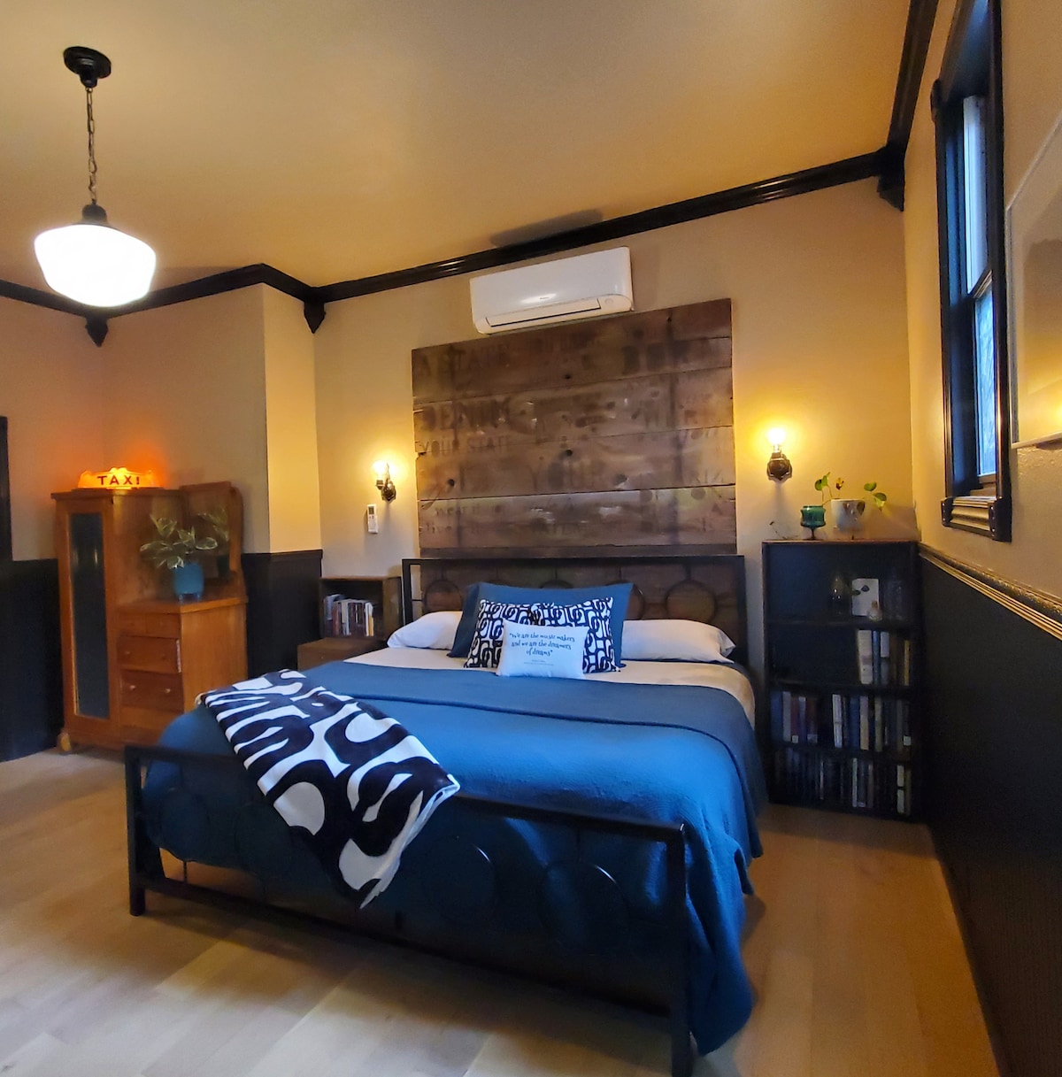 A well-appointed bedroom features a spacious bed with a patterned blanket draped across it. Vintage-style lighting fixtures are mounted on the walls, complementing the wooden accent behind the bed. A small bookshelf and a wooden cabinet provide functional storage while decorative plants add a touch of greenery.