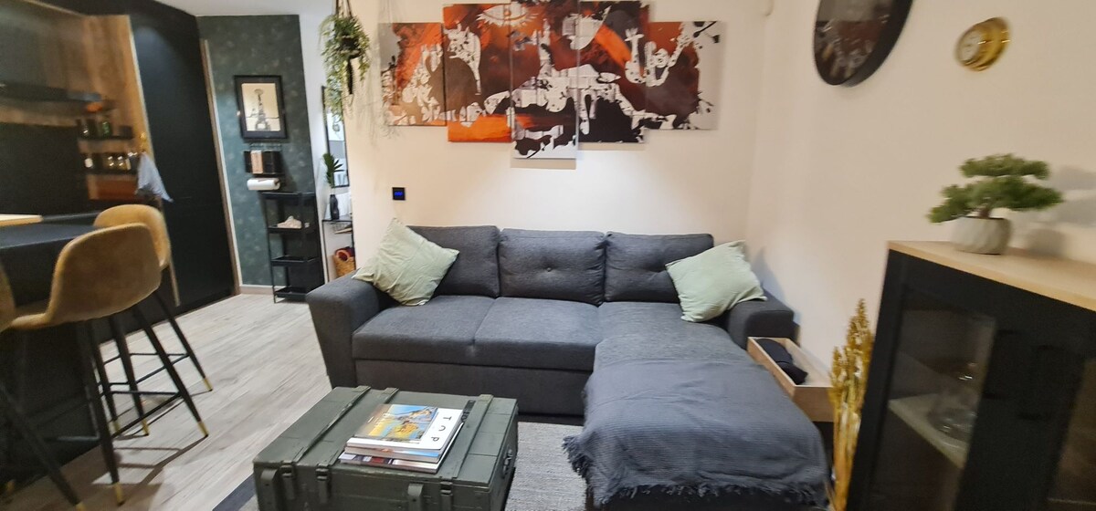 A contemporary living area showcases a dark gray sofa adorned with light green cushions. An artistic wall display adds a splash of color above a modular coffee table. The space is complemented by a stylish bar area in the background, inviting relaxation and comfort.