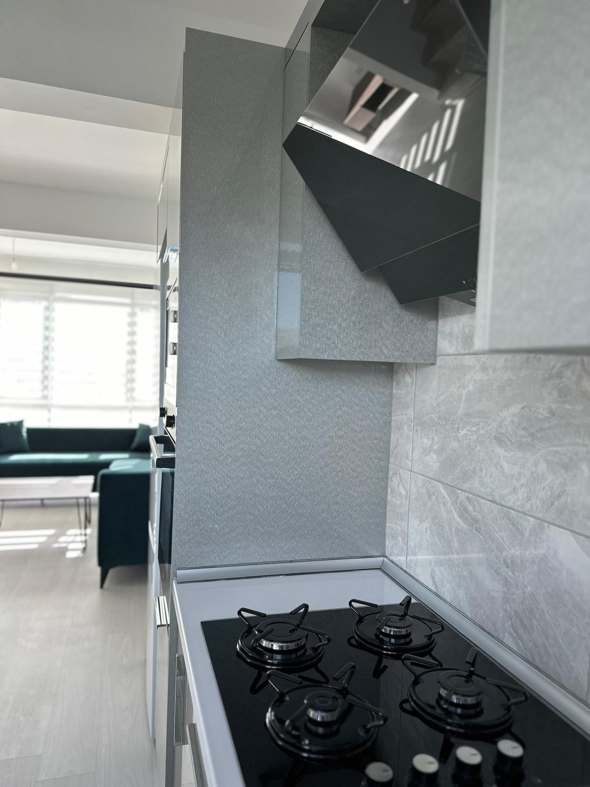 A modern kitchen with sleek cabinetry is presented alongside a gas stove featuring four burners. Natural light filters in through a nearby window, illuminating the marble-patterned backsplash and sleek countertops. A comfortable seating area can be seen in the background.