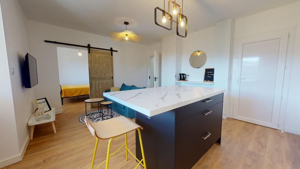 A modern open-plan kitchenette is featured with a stylish countertop and minimalist bar stools. Light wooden flooring complements the design, while a round mirror adds a contemporary touch. A sliding door leads to a sleeping area, offering additional privacy. Natural light fills the space.