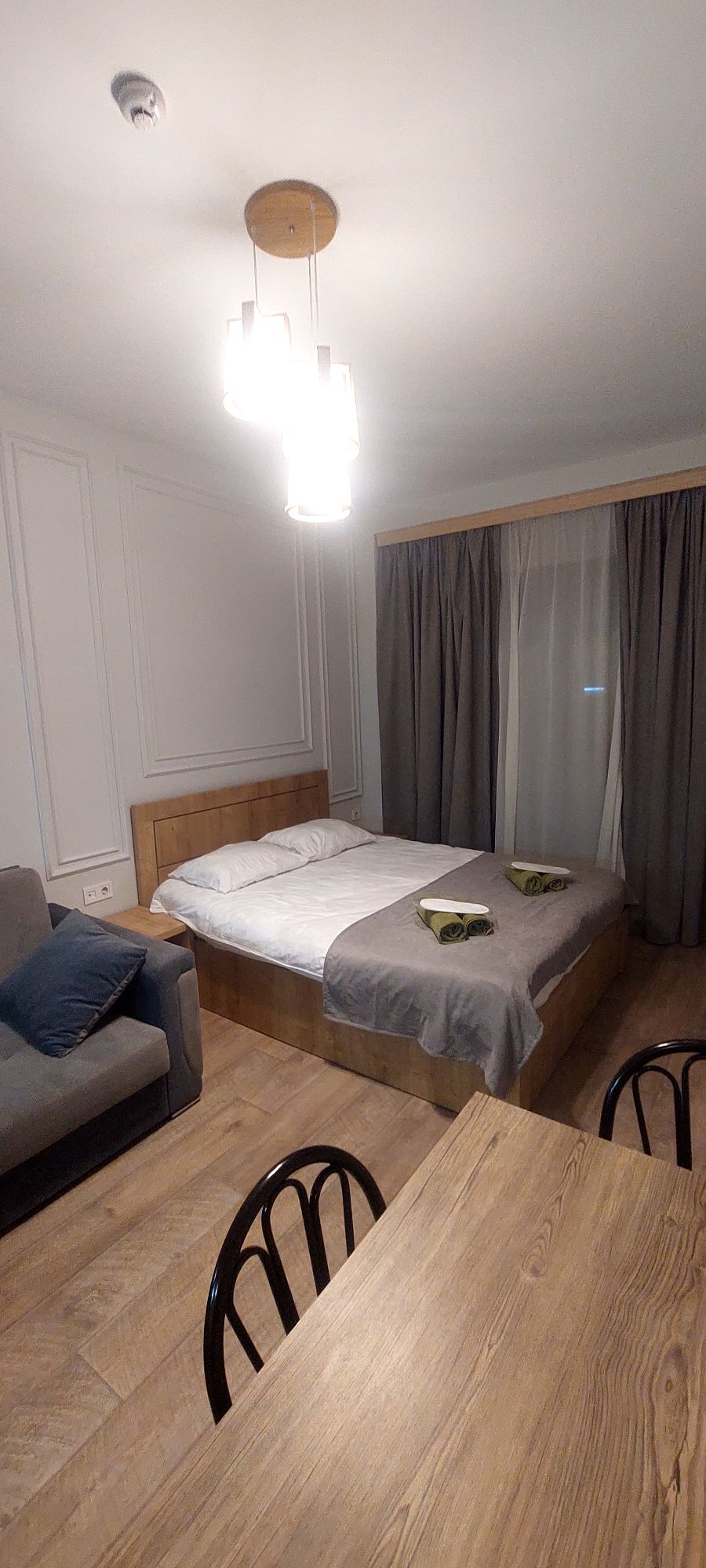 A cozy sleeping area features a spacious bed with light-colored bedding, complemented by two folded towels. A gray sofa is positioned nearby, alongside a wooden dining table with black chairs. Natural light enters through sheer curtains, enhancing the room's warm ambiance.