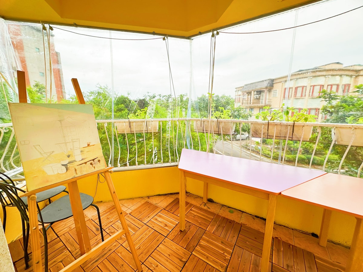 A sunlit balcony features a wooden easel holding an art piece, alongside two tables with colorful surfaces. A wooden floor adds warmth, while transparent coverings allow for views of surrounding greenery and nearby buildings.