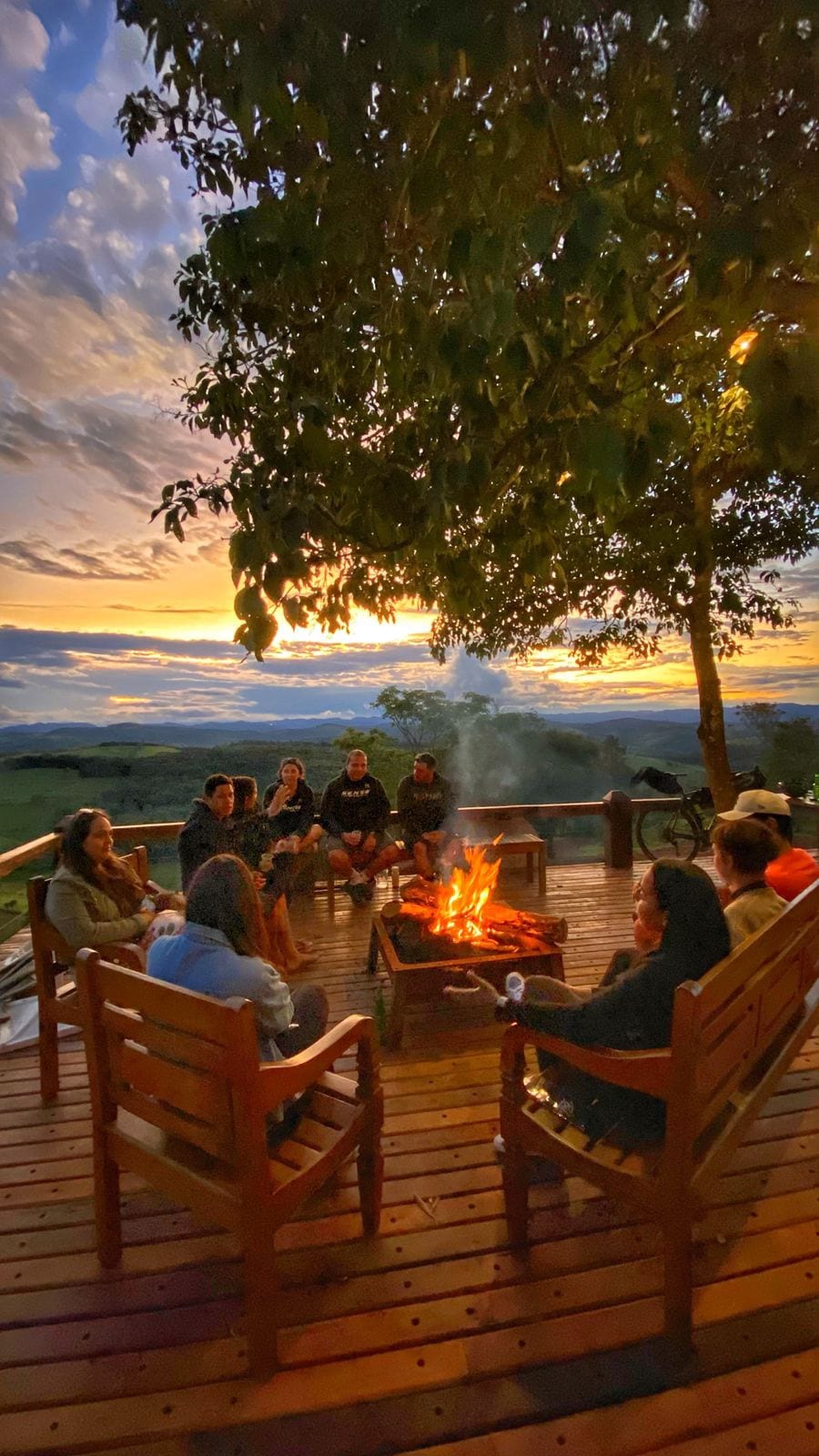 A wooden deck features a circular seating arrangement around a central fire pit, creating a communal atmosphere. Guests are gathered, enjoying the warmth of the fire under a tree. A sunset sky with hues of orange and purple serves as a backdrop.