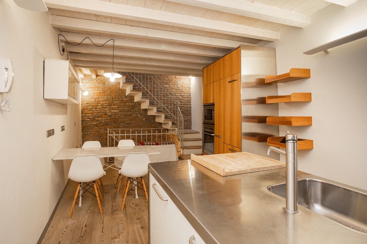 An open-plan kitchen and dining area is showcased, featuring a wooden dining table with four white chairs. A modern kitchen island with a stainless steel sink and wooden cutting board enhances the functionality. Exposed brick walls and a staircase add warmth and character to the space.