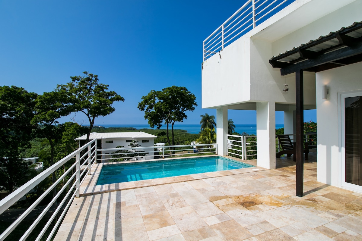 A private pool is framed by a spacious sun terrace with natural stone flooring. Lush green trees provide shade, while the stunning blue ocean and sky are visible in the background. A modern building structure showcases clean lines and expansive railing.