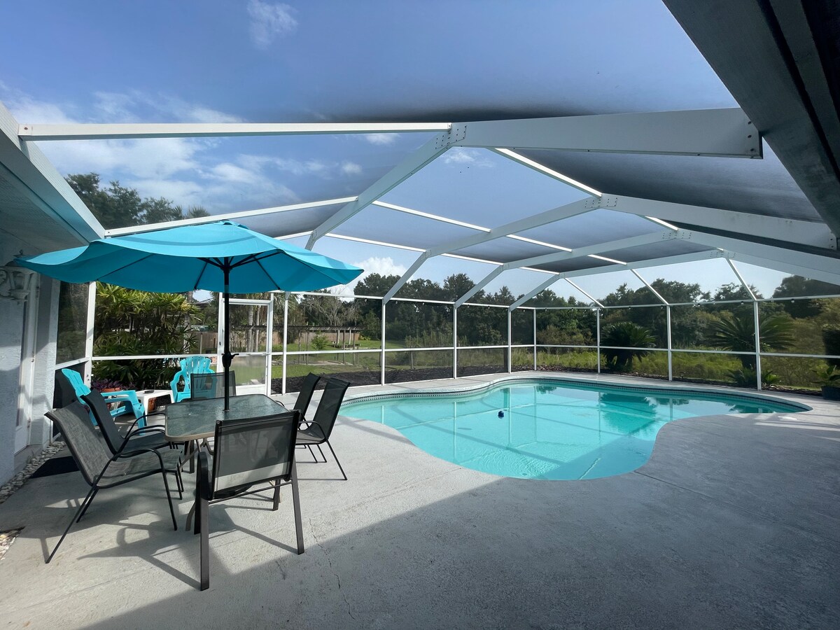 An outdoor pool area is enclosed by a screened-in lanai, featuring a clear blue pool along with an umbrella-shaded table and six chairs. Sunlight creates a bright atmosphere, while a lush green background is visible beyond the enclosure.